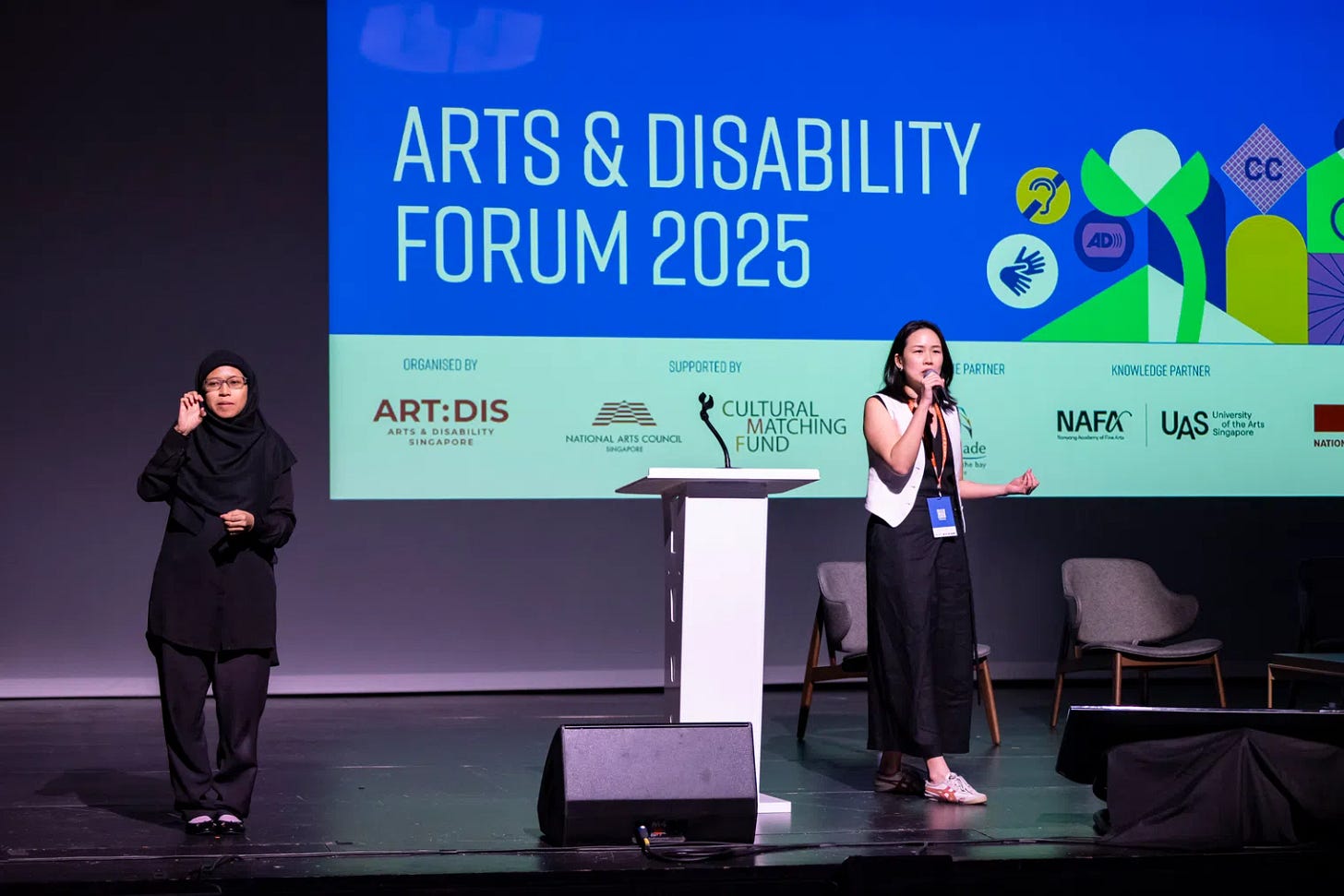 A photo of Angela Tan, executive director of Art:Dis, on a stage speaking into a handheld microphone next to a podium and a sign language interpreter. On a large projector screen behind them is a blue slide that says, Arts & Disability Forum 2025 with a digital illustration of a garden flowering into various access symbols and a bunch of organizational logos at the bottom.