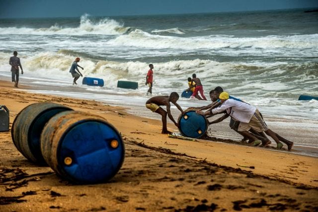 Four young men push a barrel of oil up a sandy beach