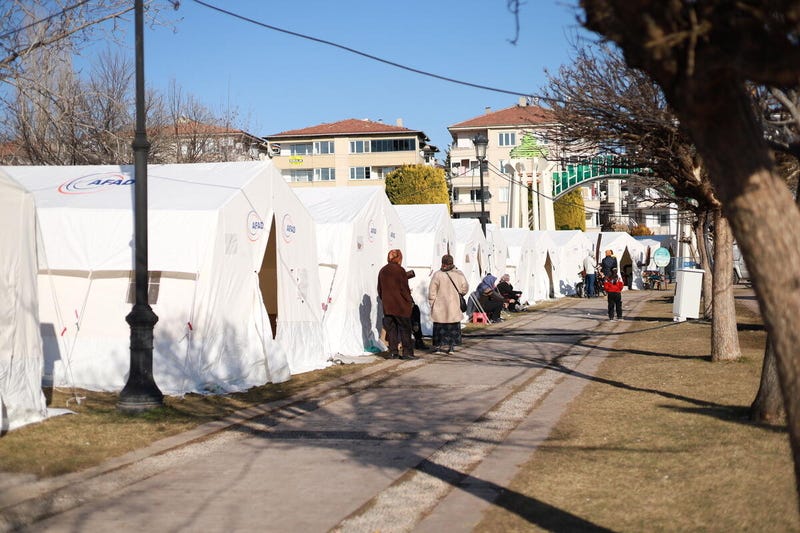 People stand and sit in front of a row of white emergency tents erected along a park walkway.