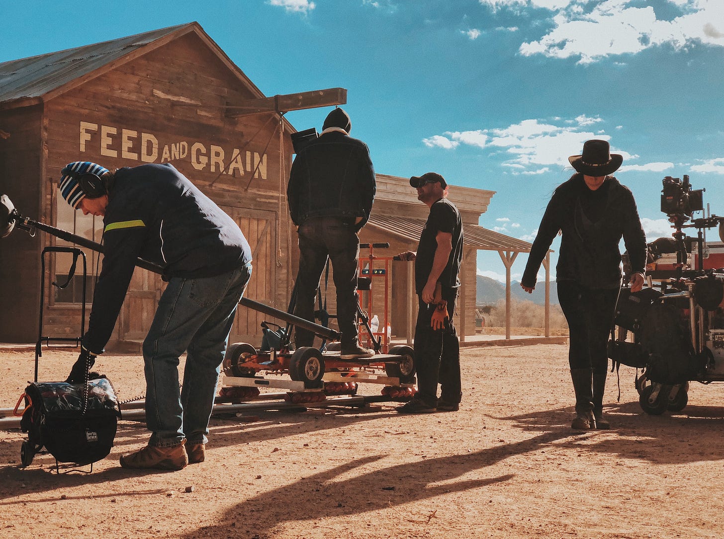 In den Kulissen einer Western-Stadt arbeitet eine Film-Crew mit ihrer Ausrüstung und bereitet eine Szene vor. Im Hintergrund blauer Himmel.