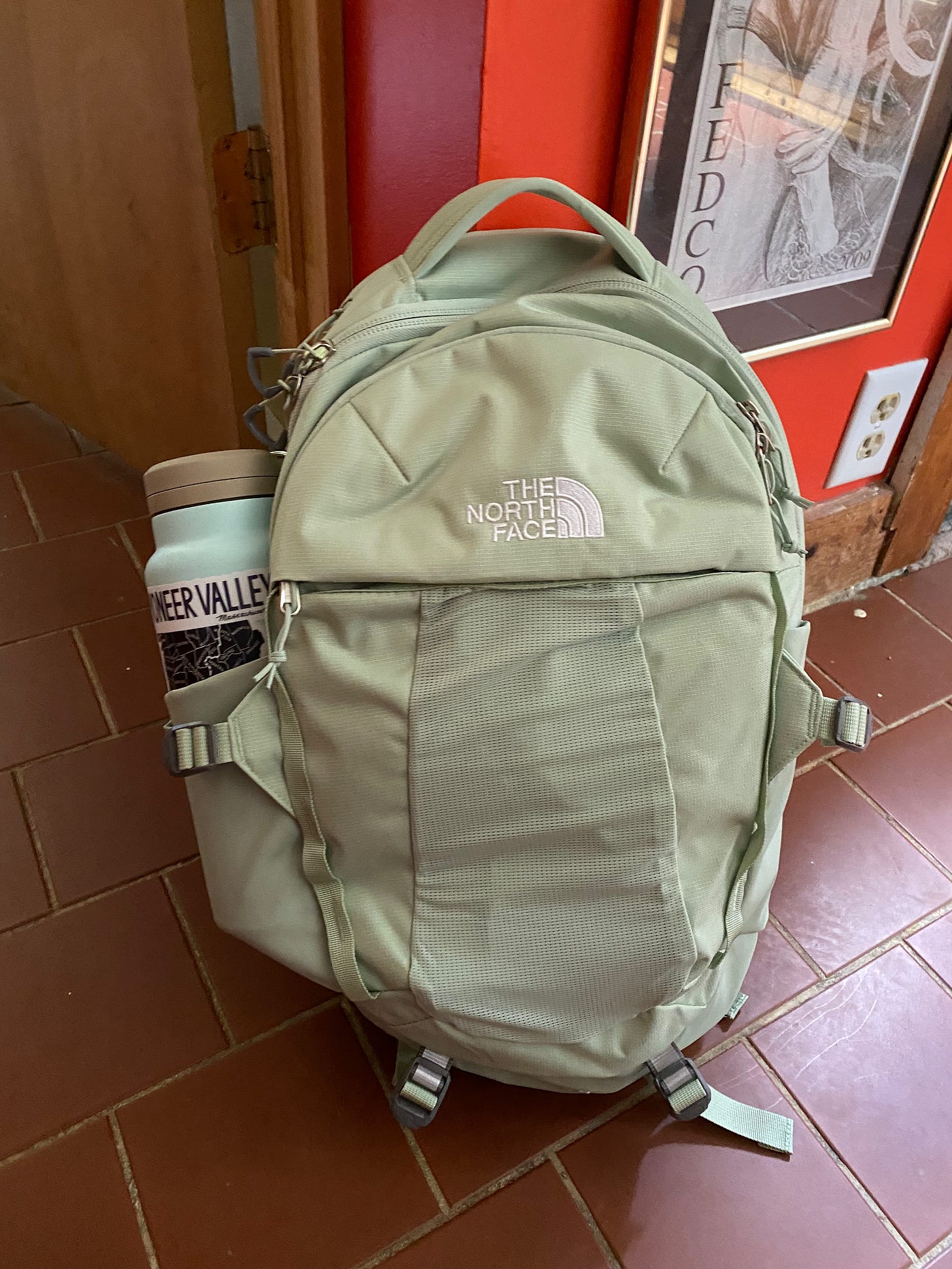 A light green backpack with a water bottle tucked into the pocket on one side leaning against a red wall.