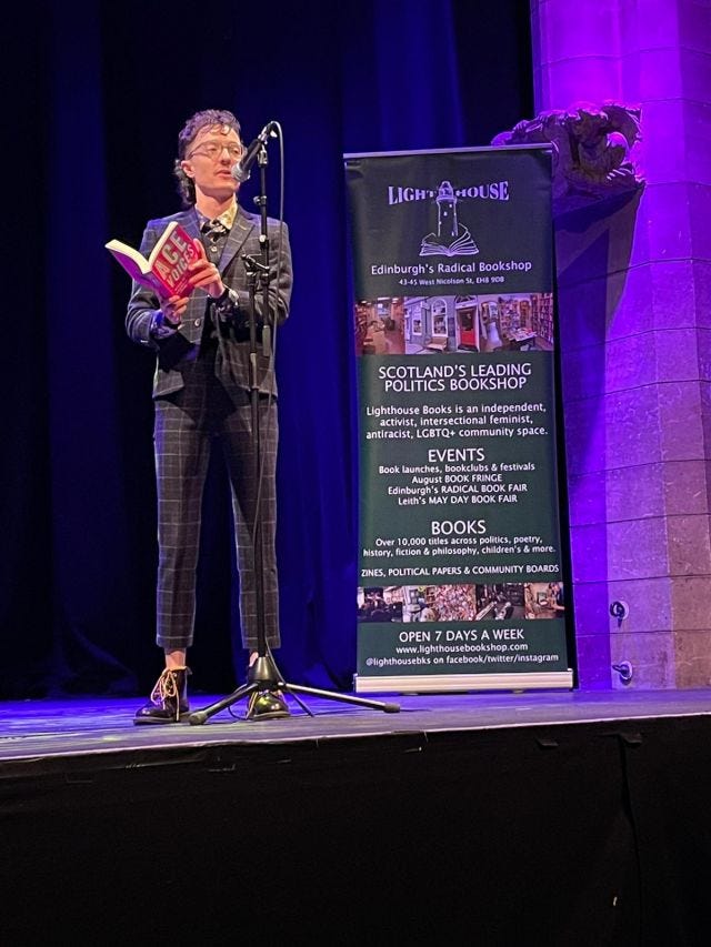 a photo of me on a purple-lit background, reading from my book Ace Voices. I'm wearing a charcoal suit in windowpane check that is very nice but slightly too small. 