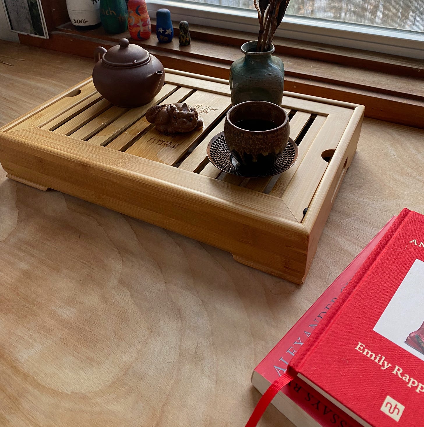 A bamboo tea tray holding a small brown teapot, tea dragon, round cup on a saucer, and small ceramic vase sits on a wooden desk under a window next to two partially-visible books.
