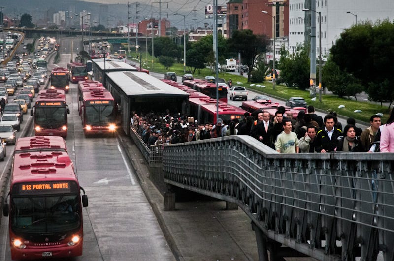 A busy Bus Rapid Transit (BRT) station in Bogotá, Colombia, with a large crowd of passengers walking on a pedestrian bridge towards the platform. Multiple red TransMilenio buses are lined up in dedicated bus lanes, efficiently moving passengers while adjacent general traffic lanes are congested with cars. The scene highlights the high capacity and effectiveness of the BRT system in contrast to private vehicle congestion.