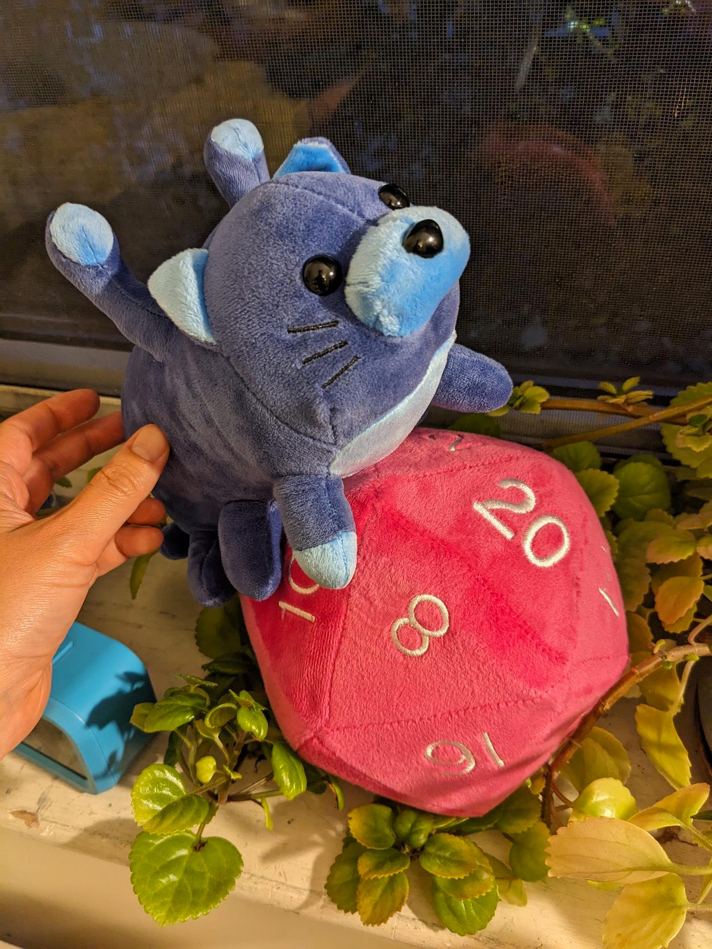 An adorable, cat-like displacer beast plushie sits atop a 20-sided pink dice plushie.