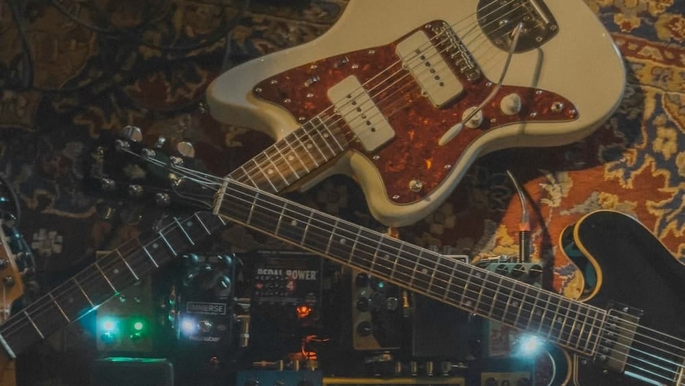 Two guitars lay on a rug next to a row of effects pedals