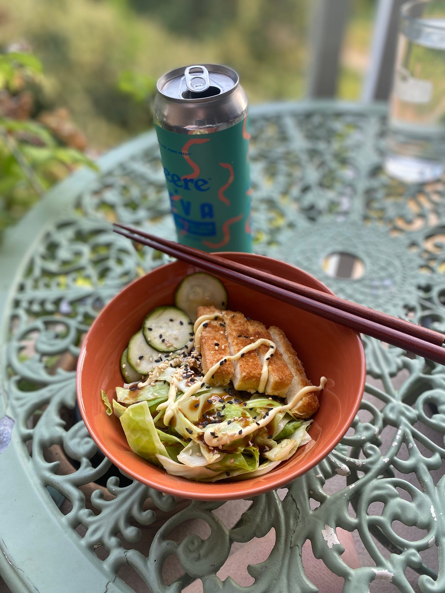 An orange bowl filled with the dish described above, the rice hidden beneath the cabbage, zucchini, and tofu. Everything is drizzled with mayo and teriyaki sauce and sprinkled with black and white sesame seeds. In the background is a blue and pink can of beer, a collab between Beere and Beva.