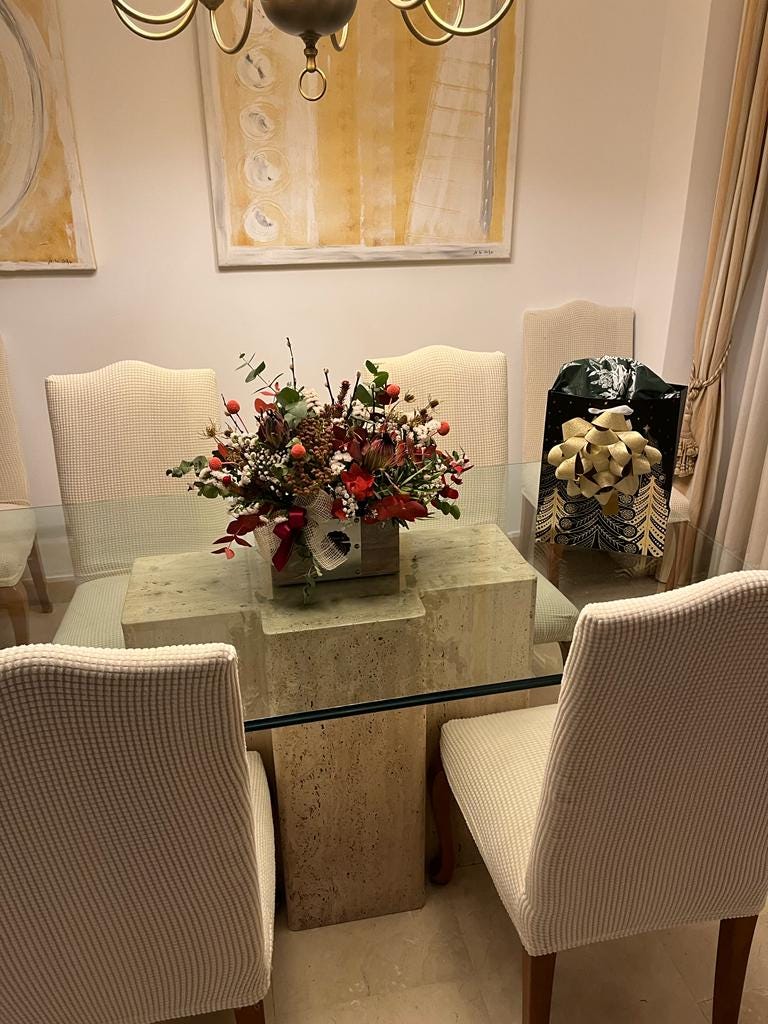 A glass dining room table with a dried floral arrangement and a Christmas gift on it; modern paintings hang on the wall behind it.