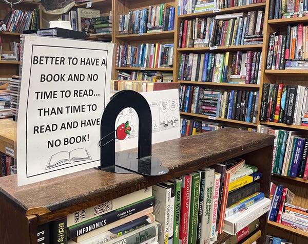 A basement bookstore with unmatched wooden book shelves overflowing with disorganized books. A homemade laminated sign reads "Better to have a book and no time to read than time to read and have no book!"