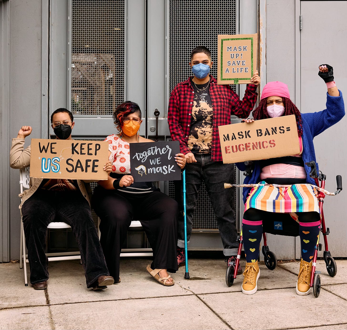 Four disabled BIPOC block a doorway with signs protesting mask bans. Everyone’s masked in a different colored KN-95.  Two people sit in chairs, one person stands with their cane, and the last person sits in their rollator with their cane across their lap. The folx on the far left and far right both have raised fists.  From left to right, the signs say, “We keep us safe,” “Together We Mask,” “Mask up! Save a life,” and “Mask bans = Eugenics.”