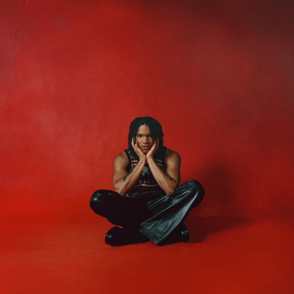 Q sits cross legged on a red floor with a red background.