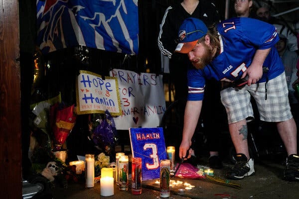 A man wearing a Buffalo Bills jersey and a Buffalo Bills hat leans down to light a candle in front of a hand-painted blue, white and red sign that reads “Hamlin, 3,” part of a memorial with other hand-painted signs and lit candles.