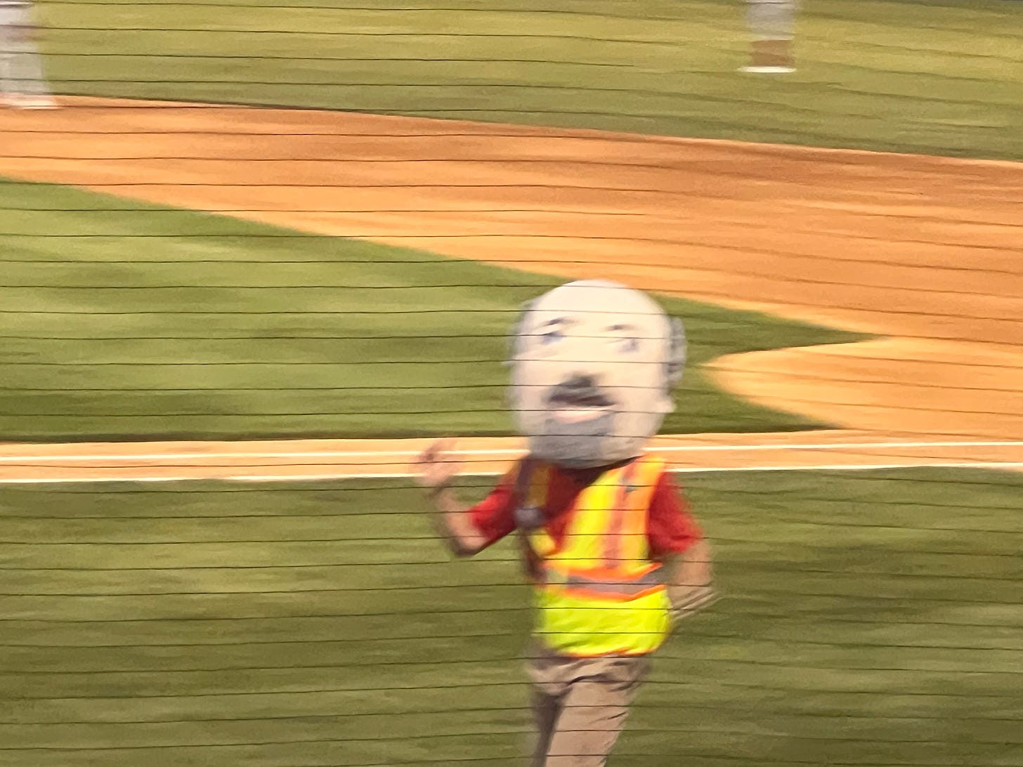 a blurry photo of a man running on a baseball field with the comically large mascot head of a goatee and balding grey hair. wearing a high vis vest