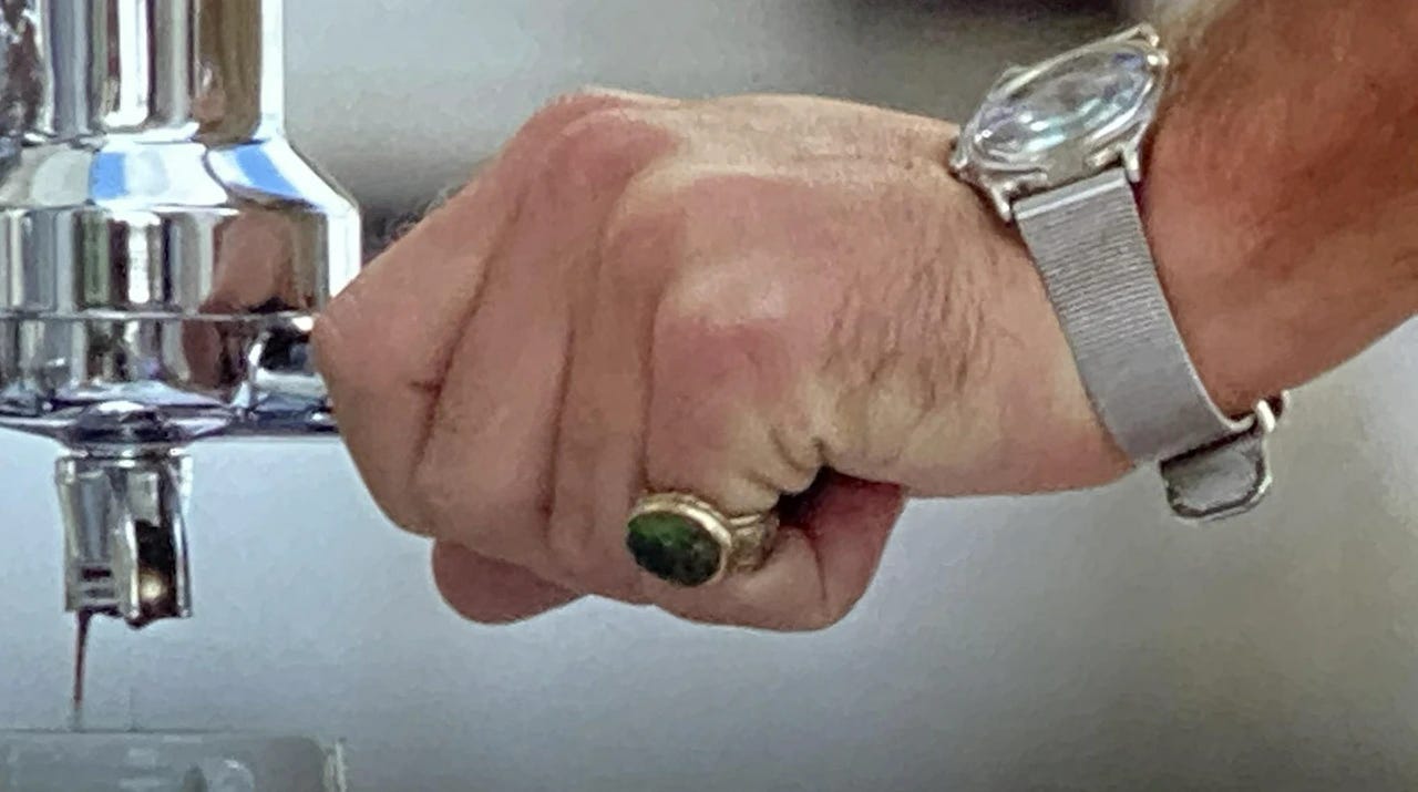 Jude Law's hand on an espresso machine handle, wearing a silver watch and chunky gold and green ring on his pinky