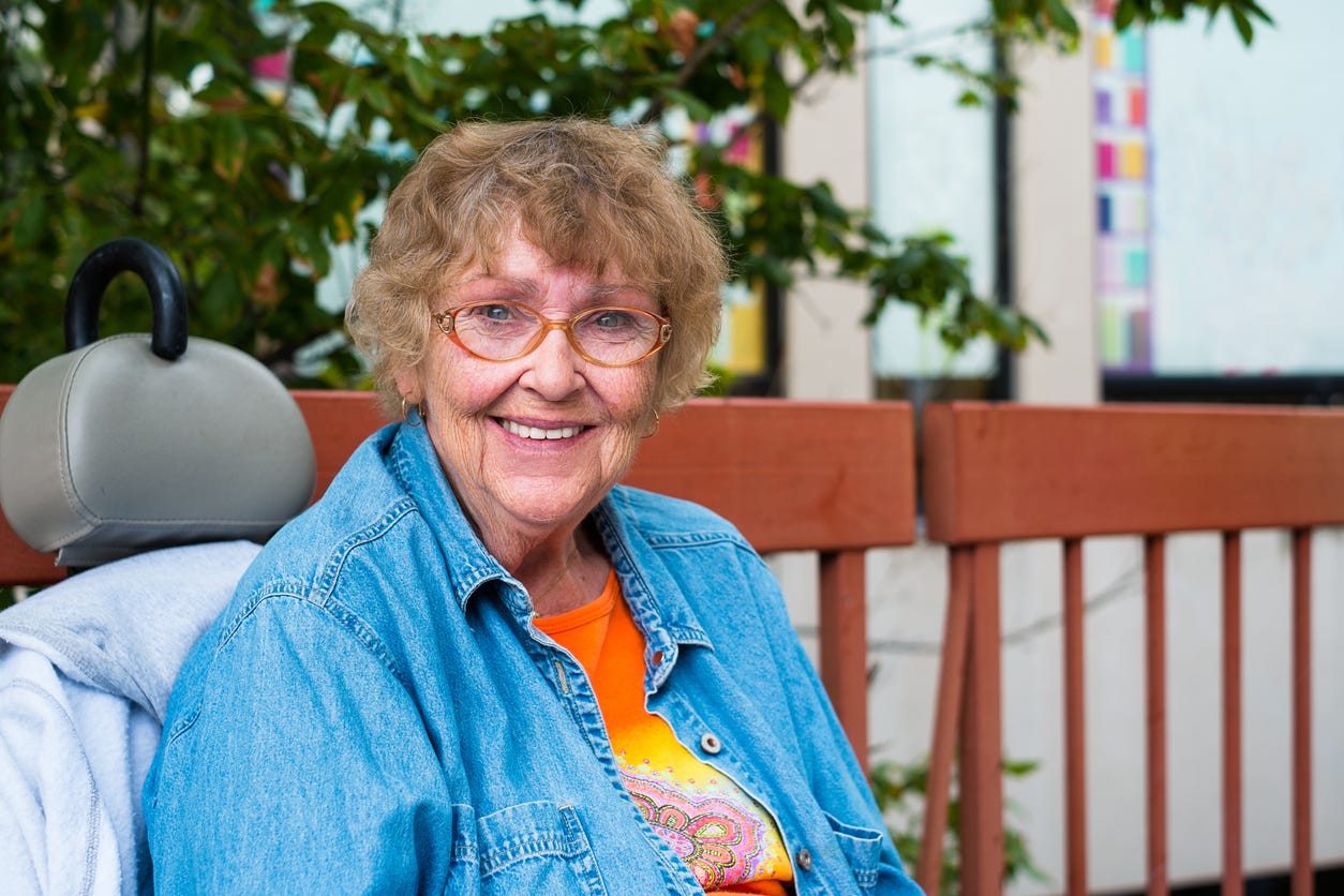 Older white woman with brown hair and orange gjasses and jeans jacket smiles pleasantly into the camera. She is sitting in her mobility scooter on a ground-level deck.