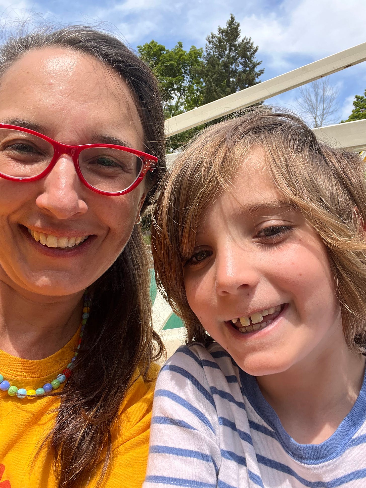 Sonya with long dark hair with gray in red glasses and a colorful shirt and necklace, next to Nathaniel who has longish hair and an impish grin, outside with trees in the background