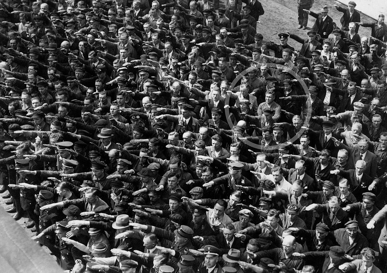 Schwarz-Weiß-Bild einer Menschenmenge (ca. 20 dichte Reihen, hauptsächlich Männer), in die linke untere Bildecke orientiert den Hitlergruß zeigend. Mitten im oberen rechten Quadranten des Bildes steht ein einzelner Mann mit verschränkten Armen, auf dem Foto durch einen grauen Kreis hervorgehoben.