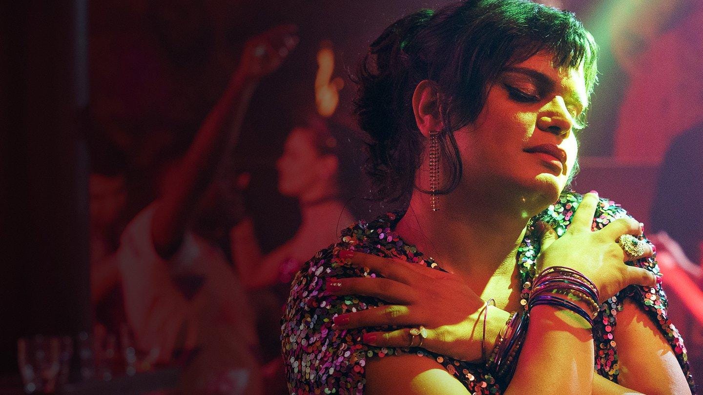 A beautiful, colorfully adorned Pakistani femme-presenting nonbinary person is on a rose-hue-lit dance floor. Their eyes are closed and their arms are crossed over their chest, in self embrace, as they seem to be dancing with themself.