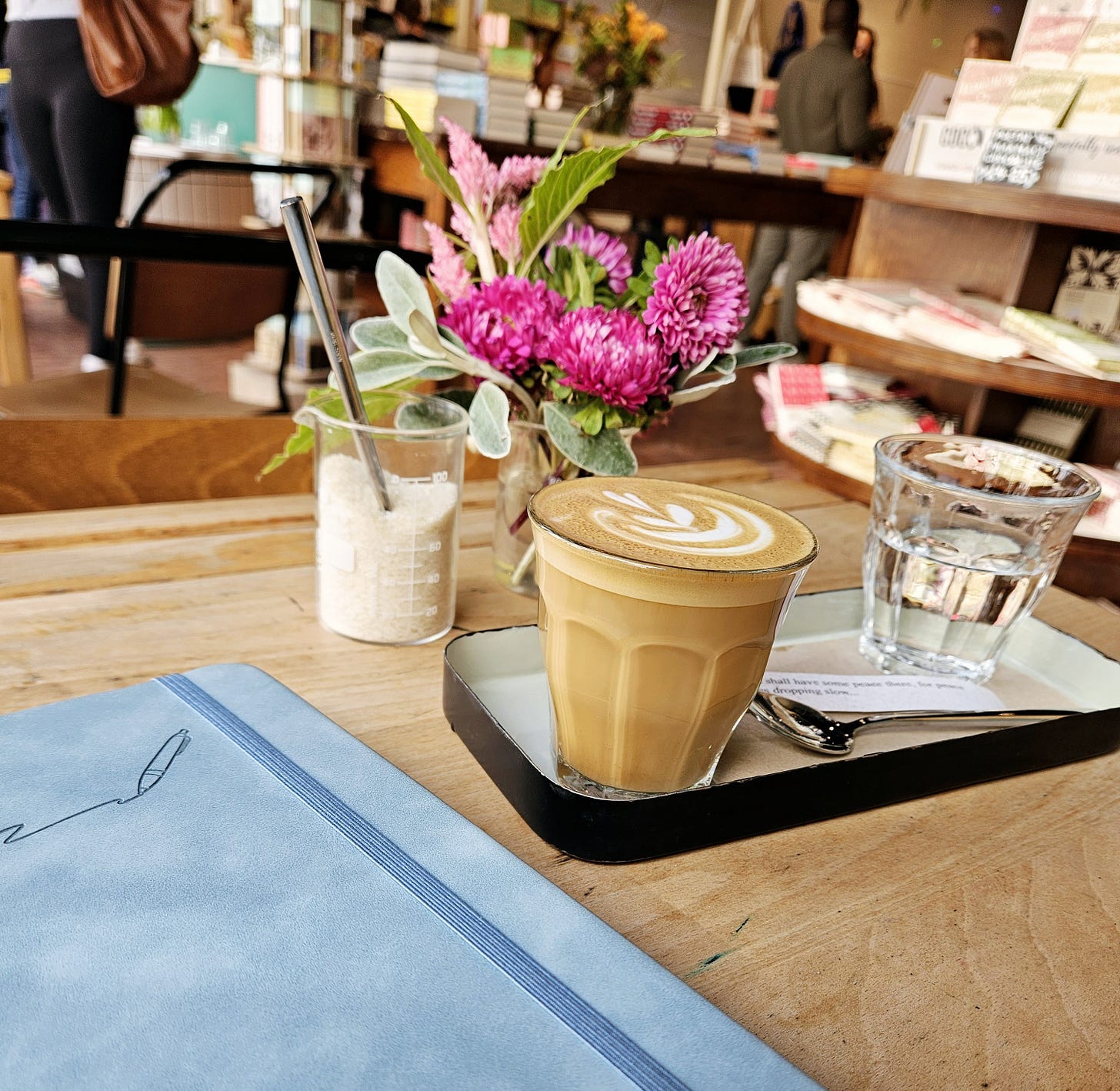 The image shows a cozy café setting. In the foreground, there is a wooden table with a black tray on it. On the tray, there is a glass of latte with a beautiful latte art design on top, a glass of water, and a spoon. Next to the tray, there is a small vase with vibrant pink flowers and green leaves. In the background, there are shelves with books and other items, and a couple of people standing, giving the place a warm and inviting atmosphere.