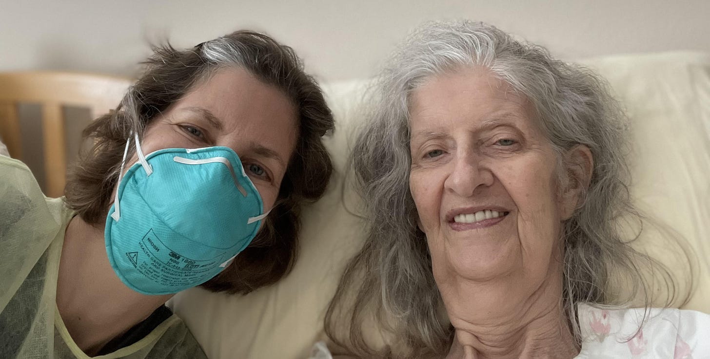 An elderly woman lies in bed near a middle-aged woman wearing a PPE mask