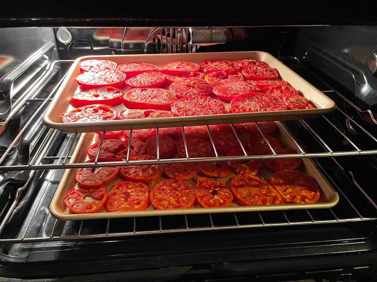 Two sheet pans full of sliced tomatoes on the racks of an open oven.