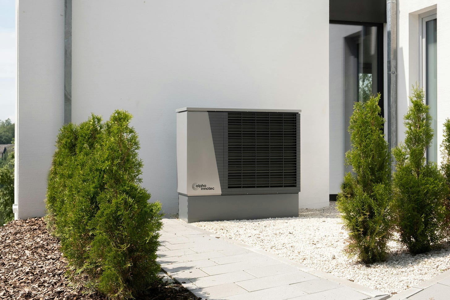 A photo of a grey, rectangular heat pump installed against the white-rendered wall of a building. Green garden shrubs and stony paths are visible in the foreground.