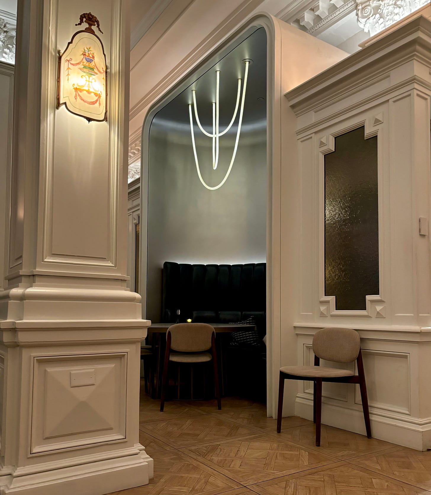 A table in a grand white room. It is in a nook offset by columns and molding. The nook itself is modern, with three strands of sleek rope lighting hanging over the table. There is a half-circle banquette against the wall.