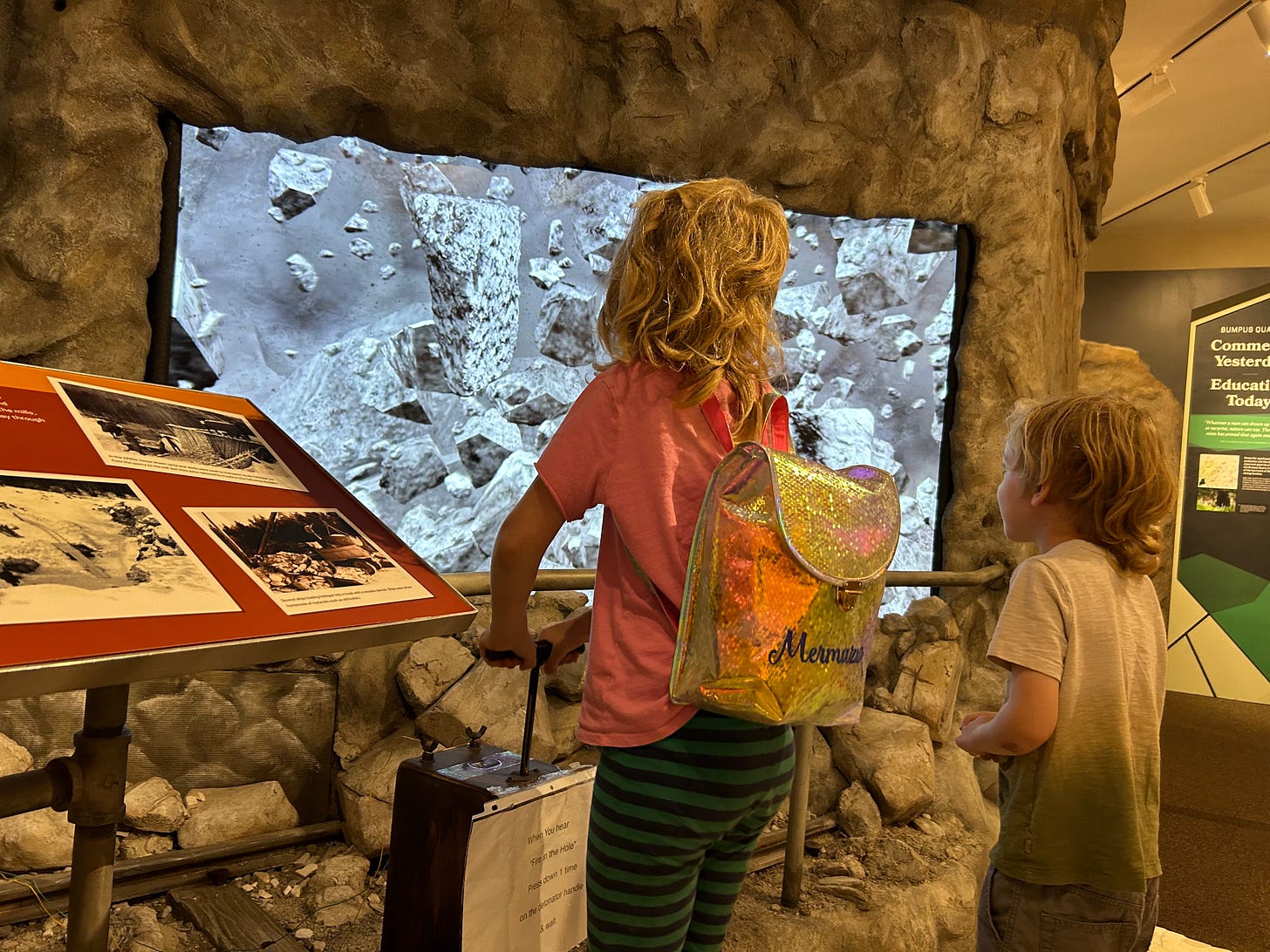 Two children with blonde hair at museum exhibit pretending to blow up part of a mineral quarry with dynamite