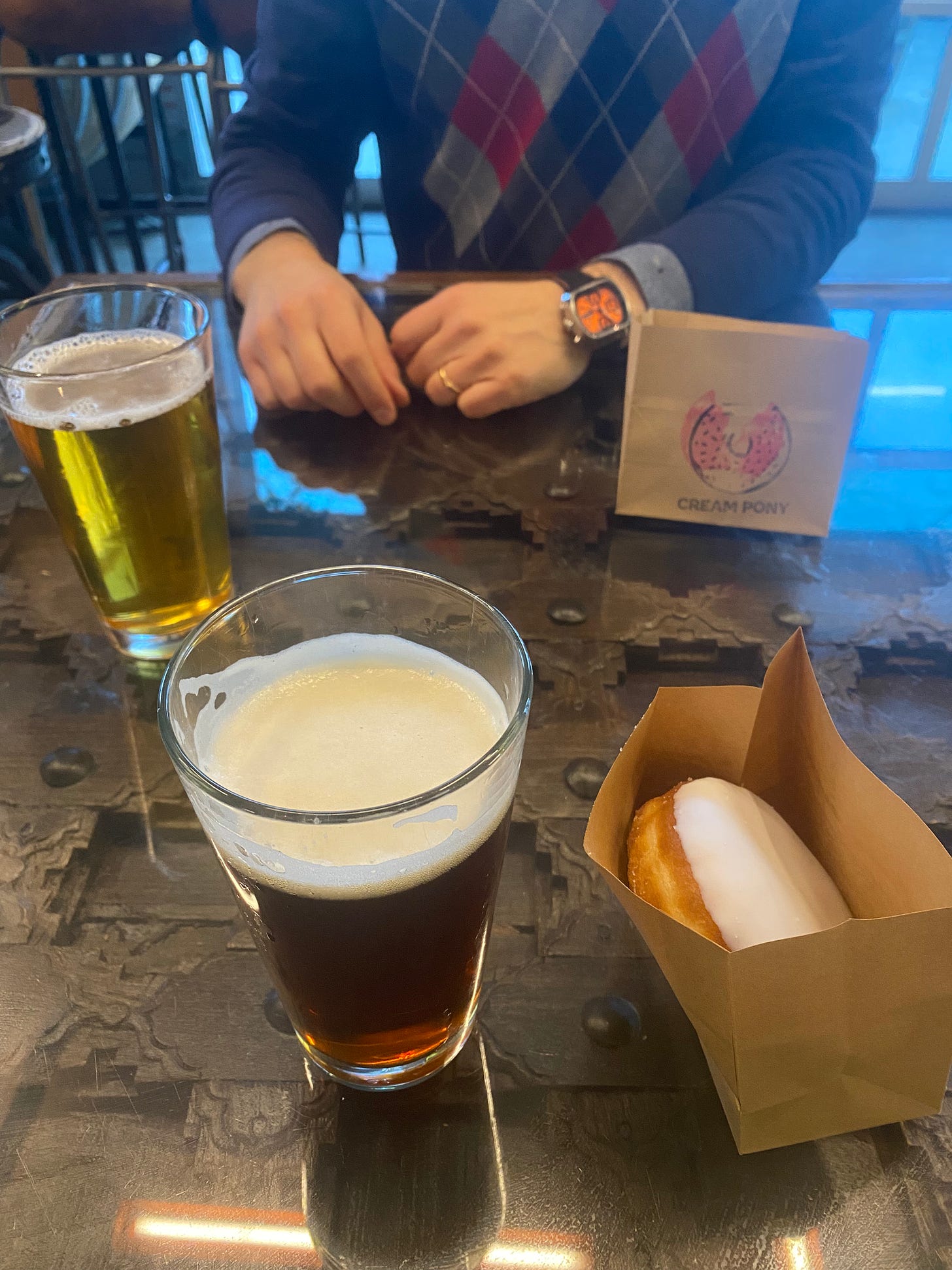 A table at House of Funk Brewing, with a glass of dark beer and a donut with white glaze in a paper bag in the foreground. In the background is a lighter beer and another paper bag with the Cream Pony logo stamped on the front. Jeff's hands are on the table between them, and he is wearing a blue argyle sweater.