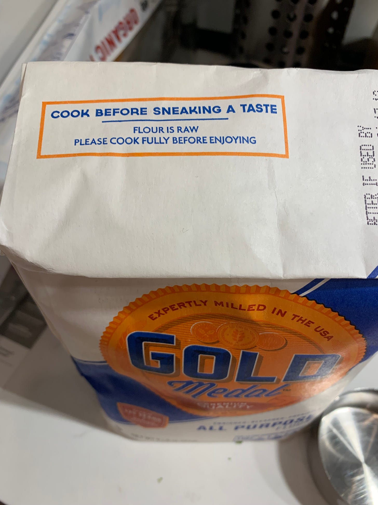 Picture shows a bag of Gold Medal All Purpose flower. On the top flap of the bag is printed "Cook before sneaking a taste. Flour is raw, please cook fully before enjoying."