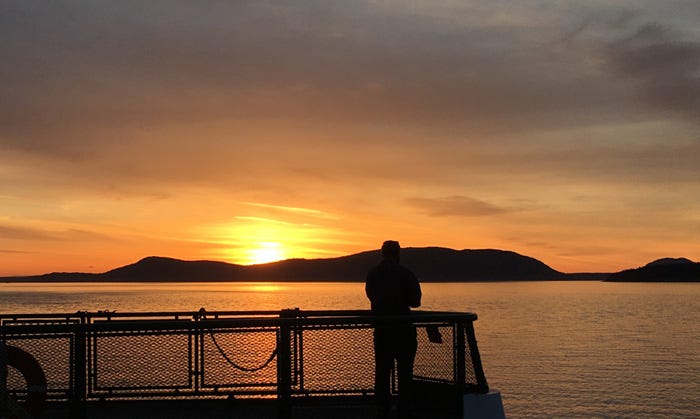 A person standing at the end of a ferry boat silhouetted against the orange sky and orange water of sunset