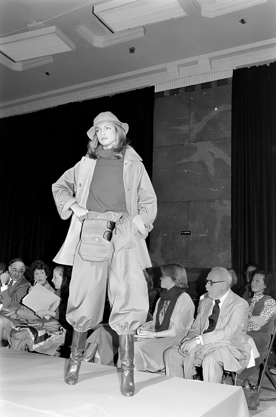 A model stands on an elevated platform with a seated audience in the background while wearing a rain hat, cowl neck sweater, oversized raincoat, wide-legged trousers cinched at the ankles with elastic, and knee-high rain boots. 