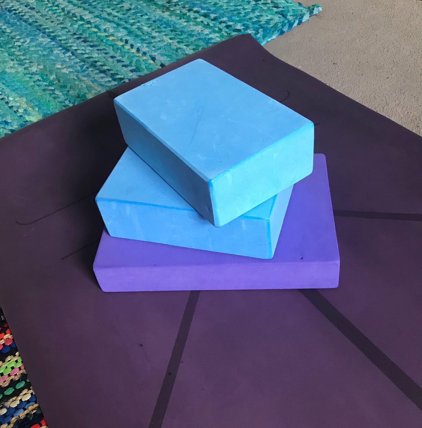 A purple yoga brick and two blue yoga blocks sitting piled on top of a dark purple yoga mat