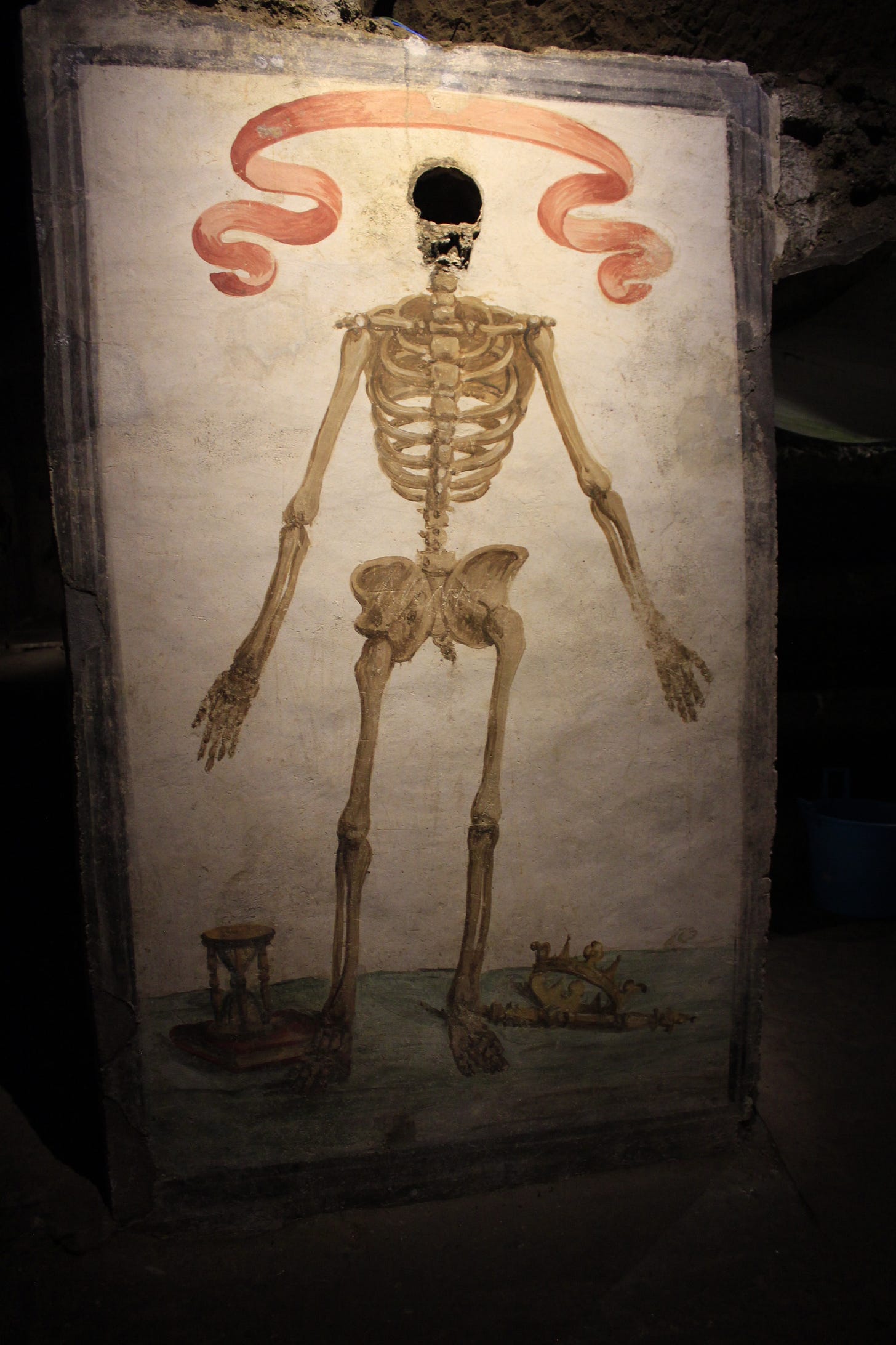 a fresco of a skeleton with a hole where a real skull was once mounted. there's a ribbon over its head and an hourglass and crown by its feet.