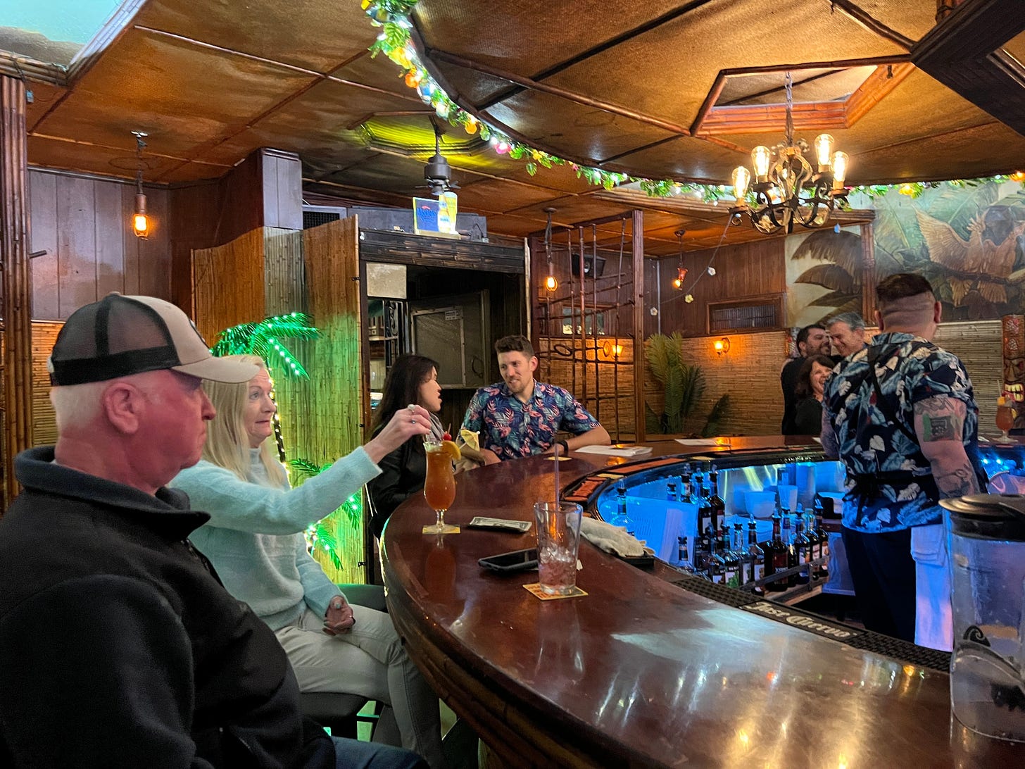 Four customer sit at a circular bar drinking cocktails and a bartender stands behind the bar in a tropical shirt.