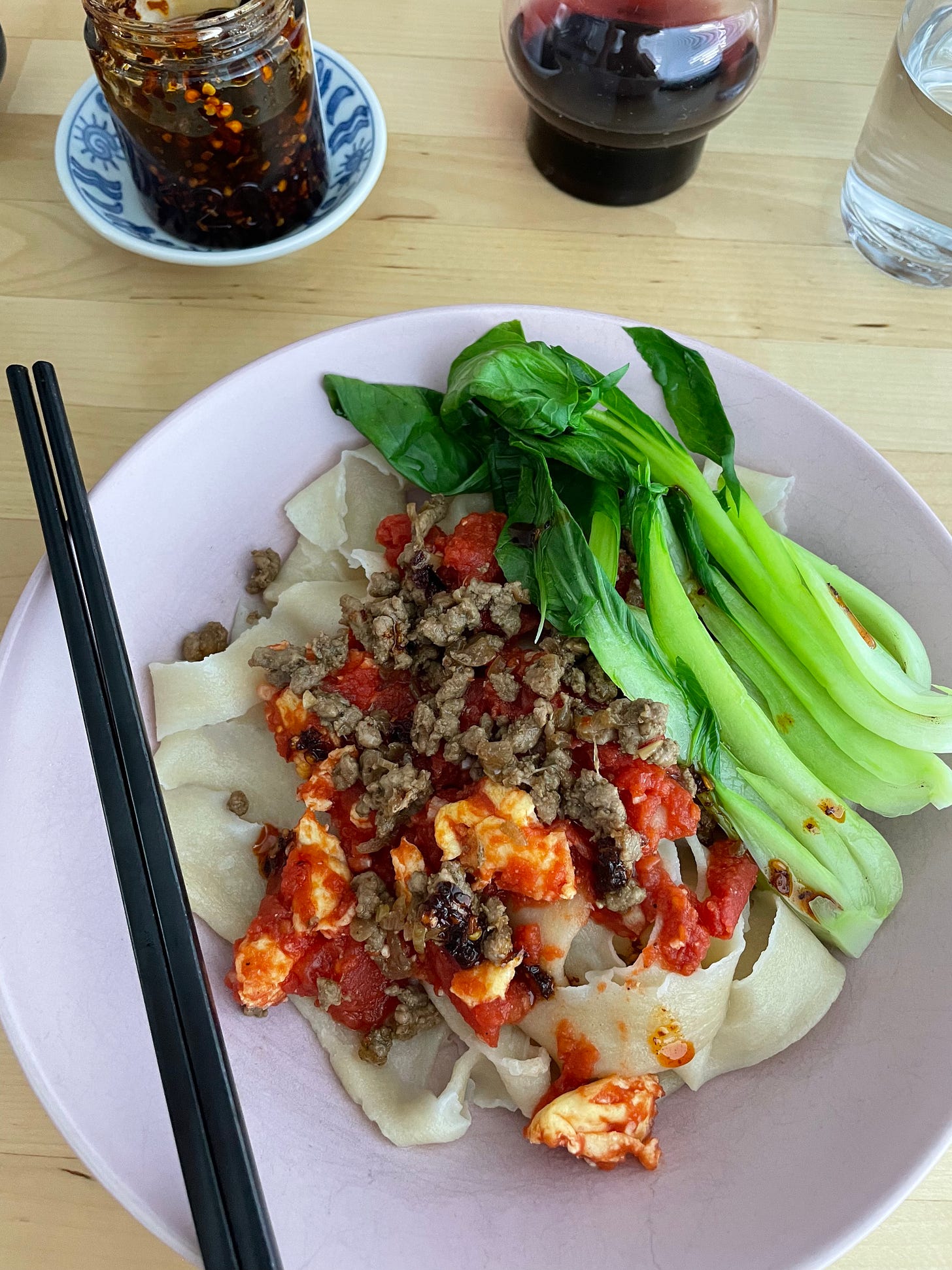 Hand ripped noodles topped with tomato and egg sauce, a side of baby bok choy and pork. Chopsticks and chilli sauce nearby.