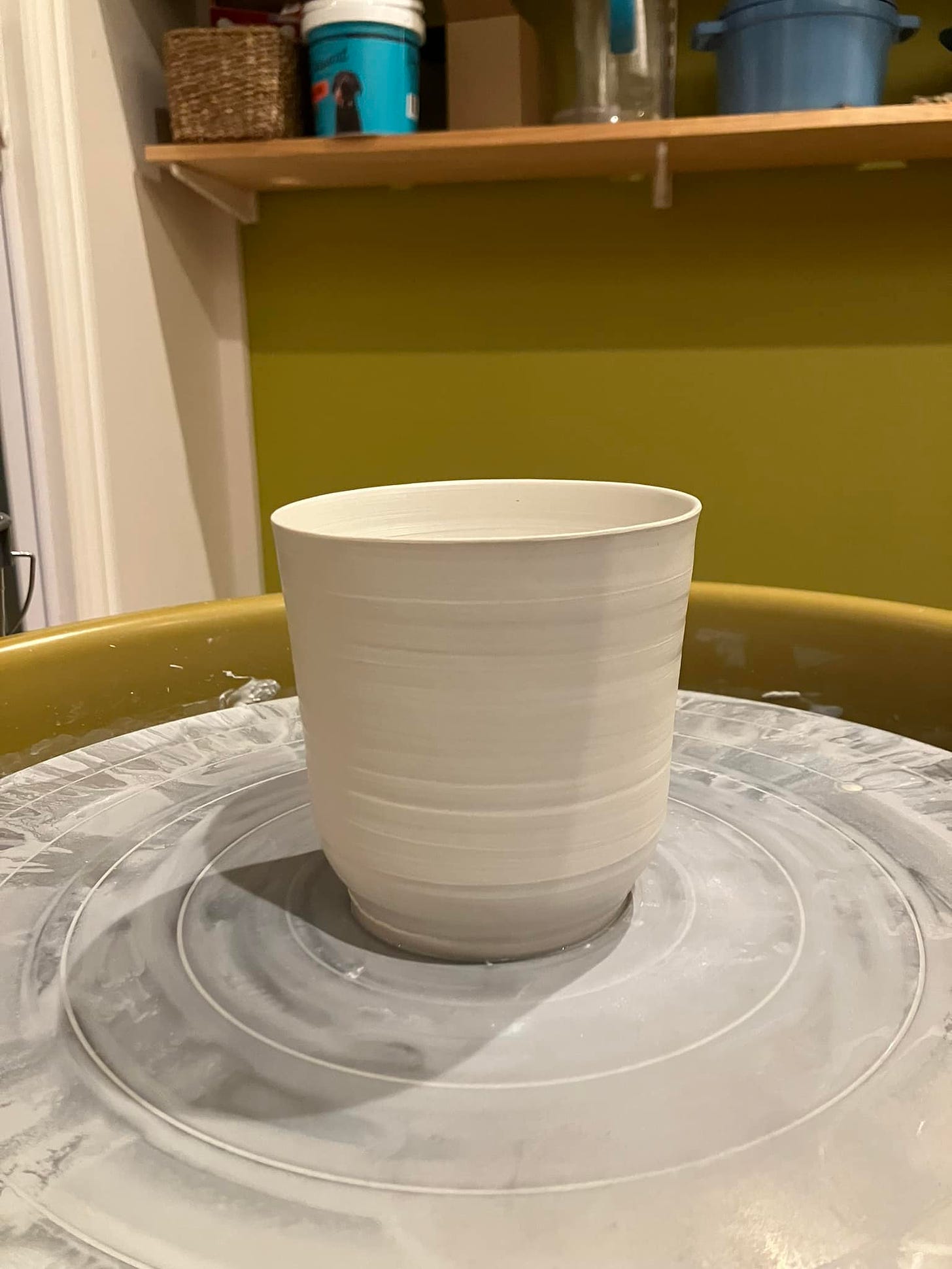 A new porcelain cup, still on the wheel. In the background is a lemongrass wall and pine shelves.