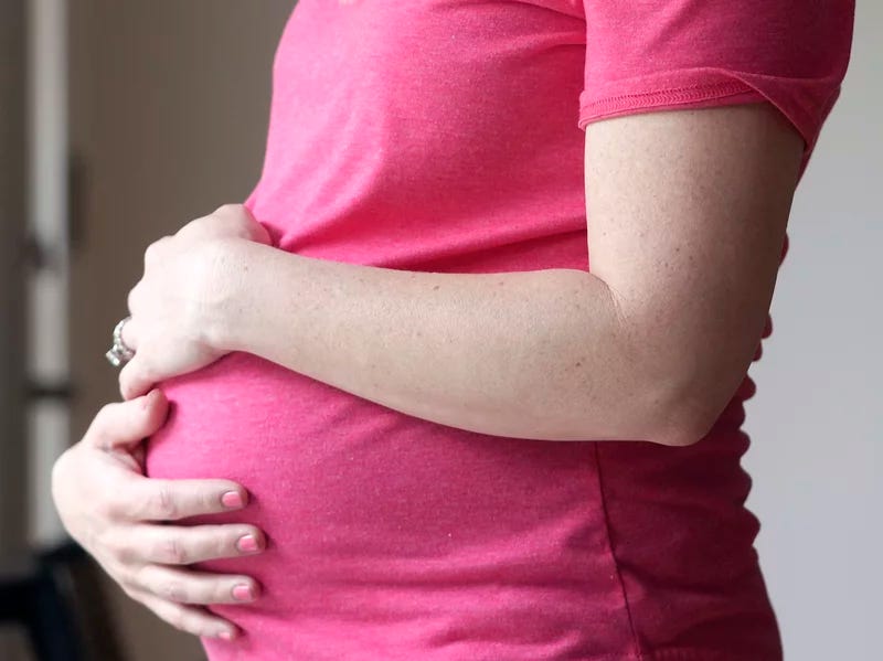 An image of a white pregnant person from the side, showing the chest down to the belly. They are wearing a pink shirt and are holding their belly with both hands.