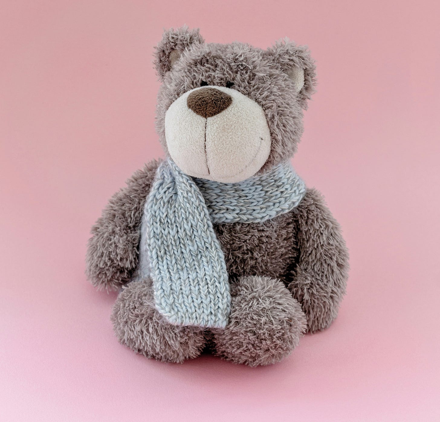 Smiling soft light brown teddy bear in a light grey scarf, against a light pink background