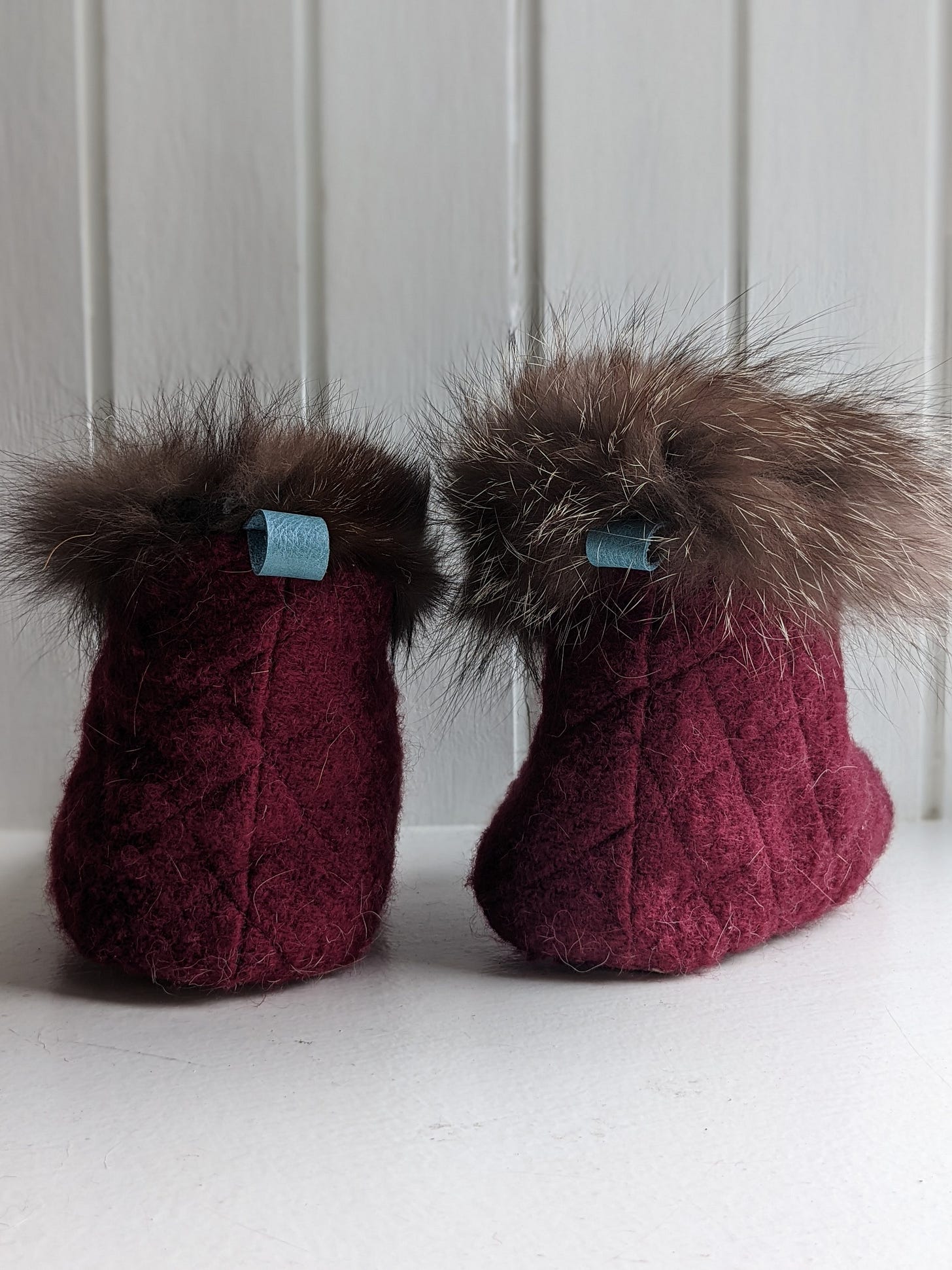 A pair of quilted wool slipper booties, from behind. They have a blue leather hanging tab and a fur cuff