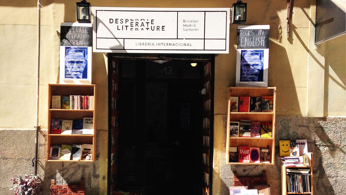 The front entrance of the bookshop, a sign for Desperate literature above the door, black on white background, two bookshelves stand on each side of the door with posters above