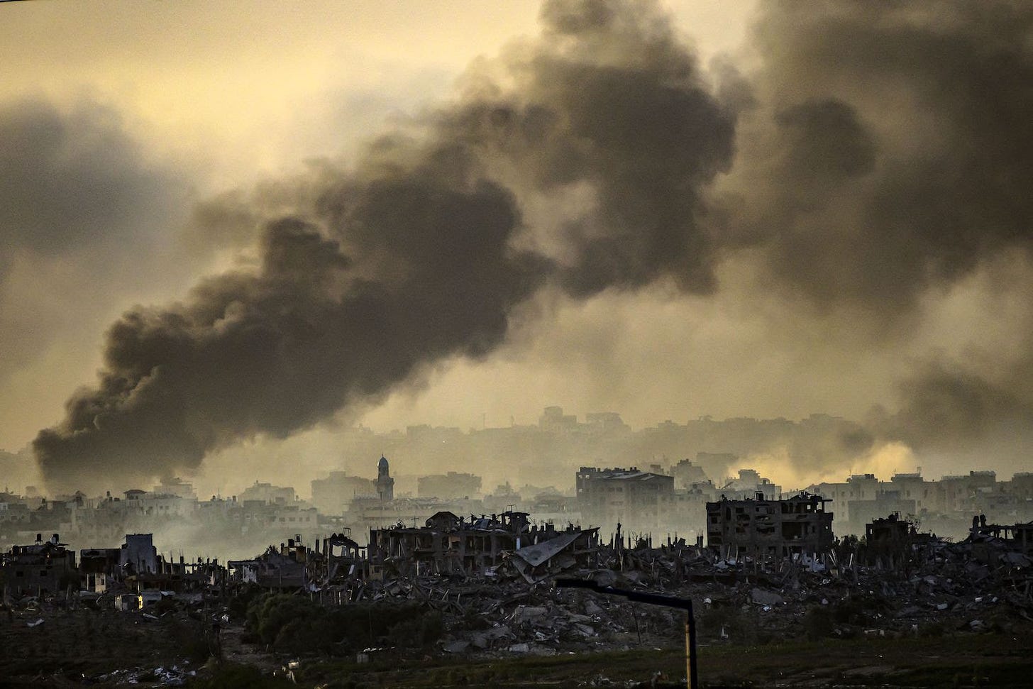 A wide shot shows a densely packed urban area of the Gaza Strip. Many of the buildings have been leveled by explosions, leaving a dark mess of rubble. Smoke billows from different locations and fills the sky overhead, overlaying the cityscape in dusty gray.