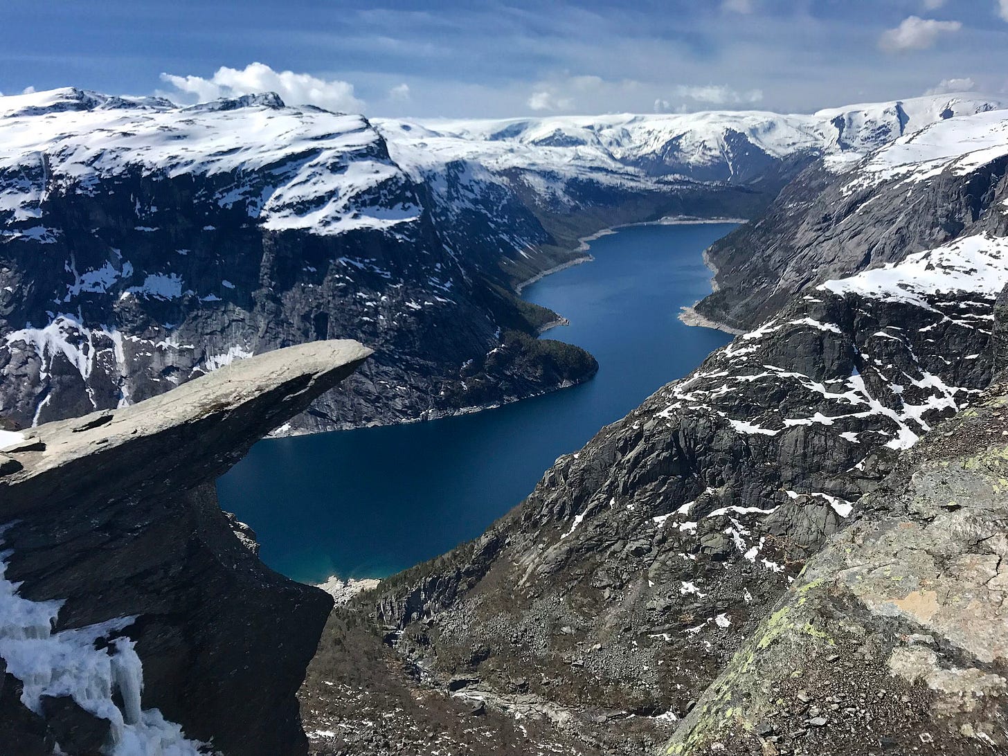 a picture of the trolltunga above a fjord, snowy mountains in the background