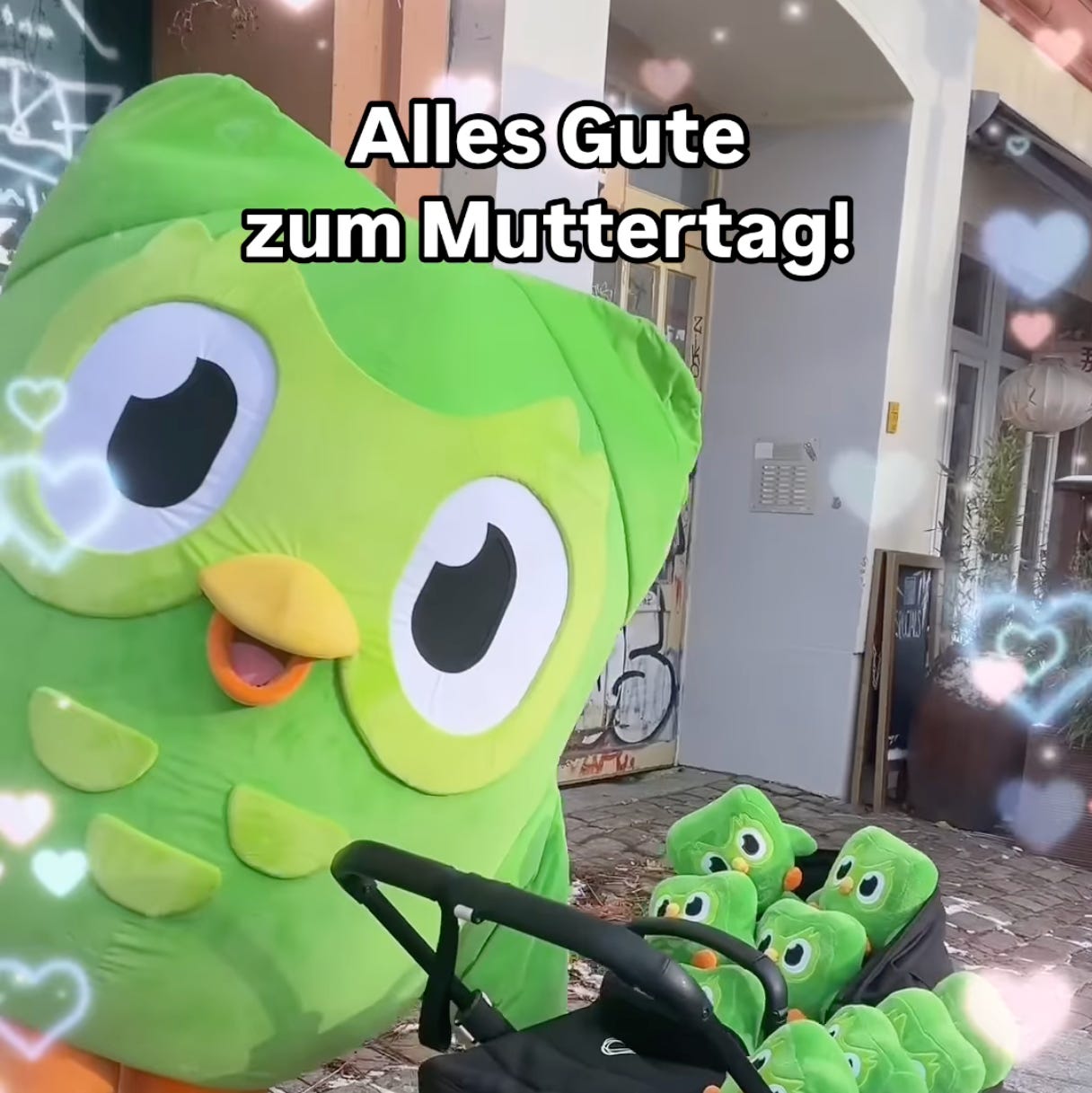 Duo, a large green owl mascot, stands over a stroller with many little baby green owls in it. The sentence "alles gute zum Muttertag" is superimposed on top.