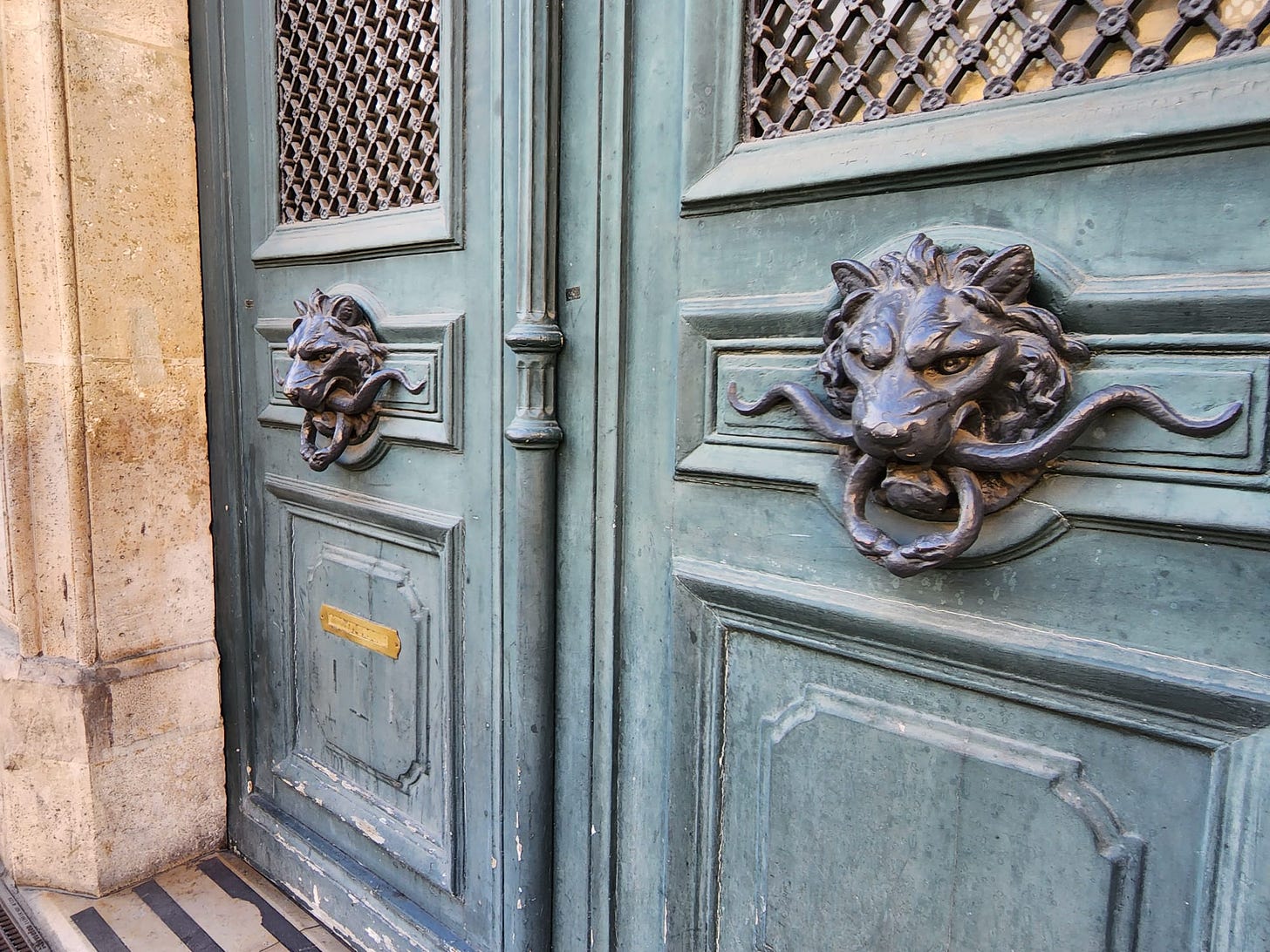 Photo of ornate and weathered light blue doors with massive lion-head knockers. 