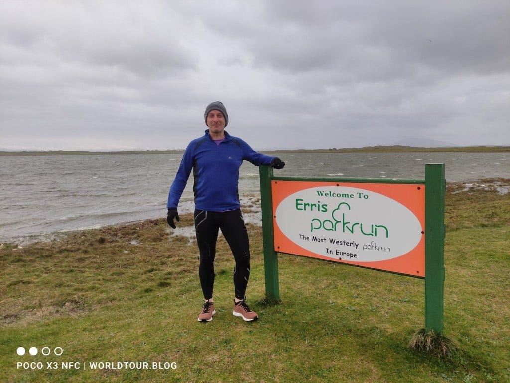 Me, pictured next to the permanent sign that marks the start of the parkrun, overlooking the lake which is lapping at the grass