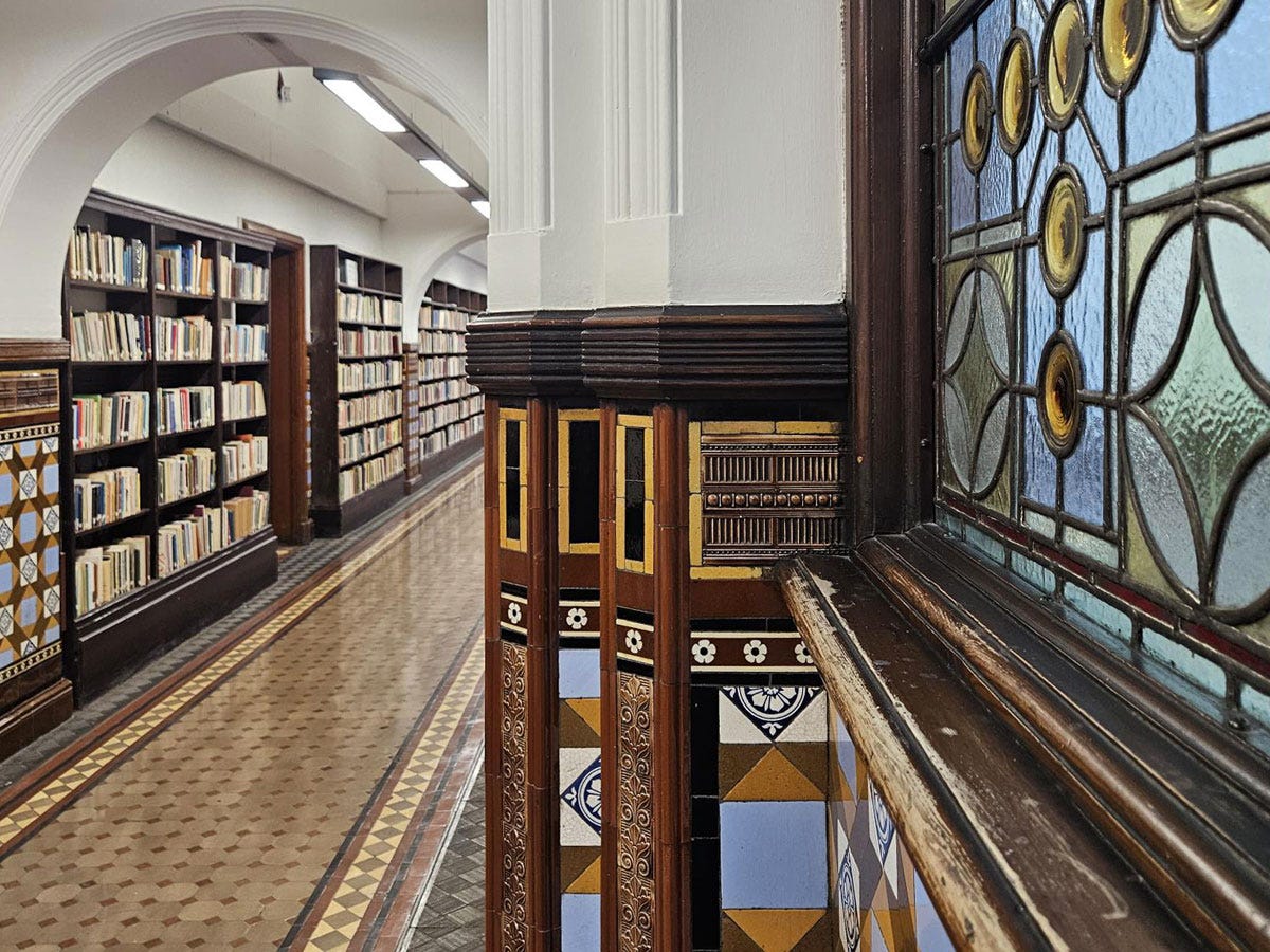 A corridor with bookcases on one side and a close-up view of a stained glass winow and tiled pillar on the other side