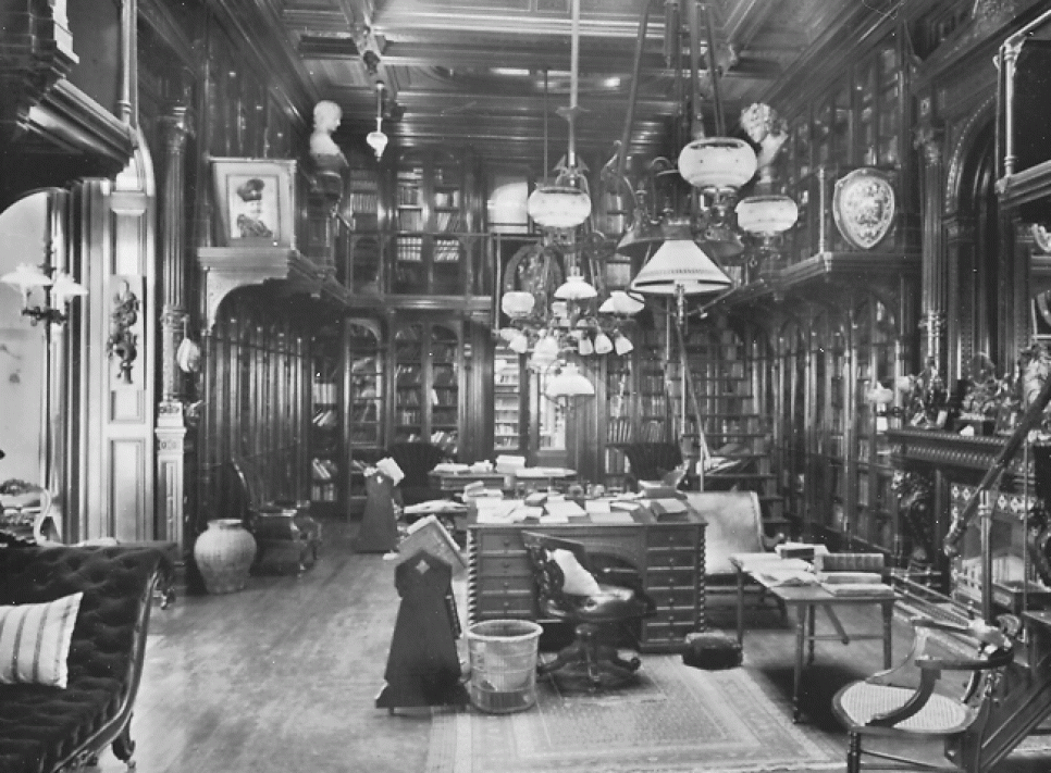 A black and white photo of a vast, ornate 19th century library with wooden bookcases, large leather-bound books, and a desk scattered with papers.