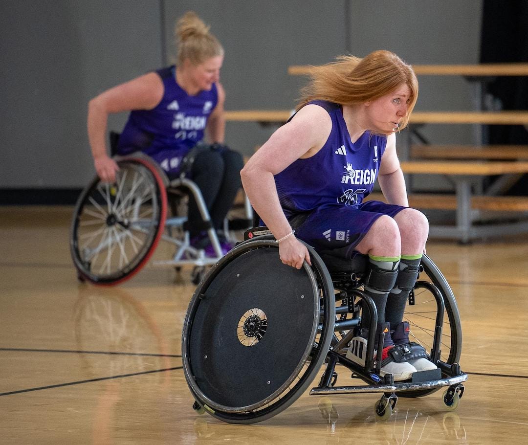 A white woman with red hair is rushing forward in a manual wheelchair. She wears a purple uniform that says REIGN with the Adidas logo.