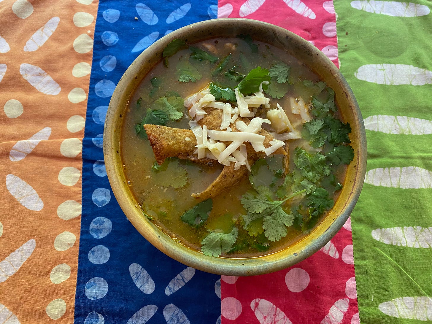 A ceramic bowl of brothy soup, topped with a pile of fried tortilla stirps, grated cheese, and fresh cilantro leaves, sits on a colorful placemat. 